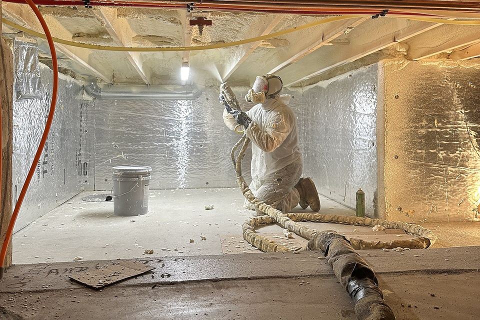 Installer spraying foam insulation onto rim joists in a crawl space lined with foil-faced boards