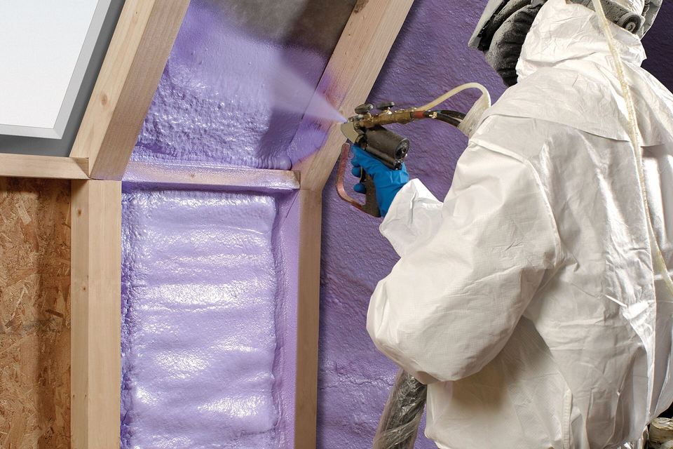 Installer in a protective suit and respirator spraying foam insulation into open wall stud cavities